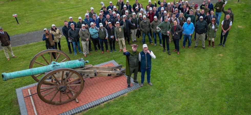 A large group of people waving to a drone next to an ornamental cannon.